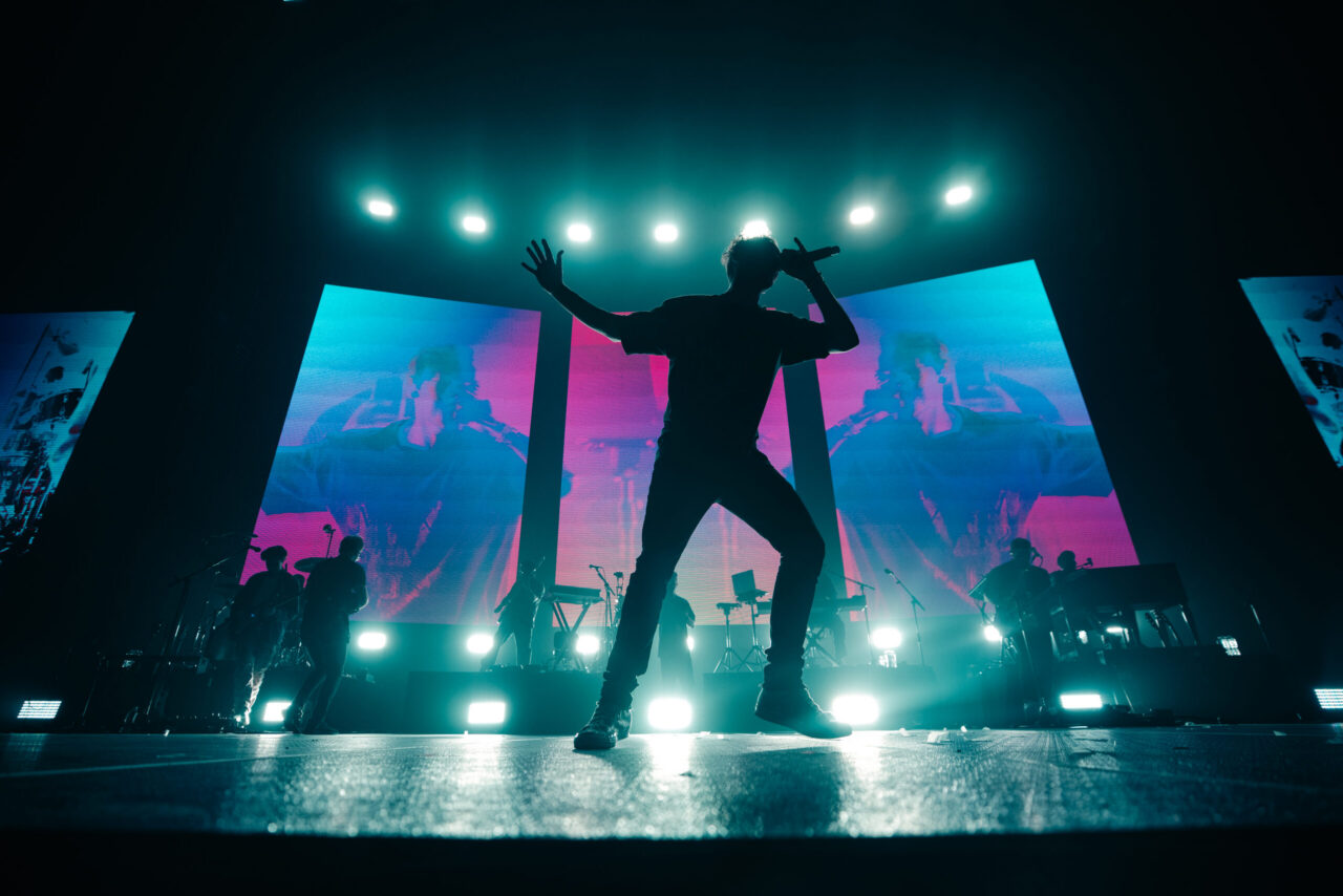 Singer rocks the stage, backlit by bold turquoise and pink lights, with bandmates and wild visuals on big screens behind.
