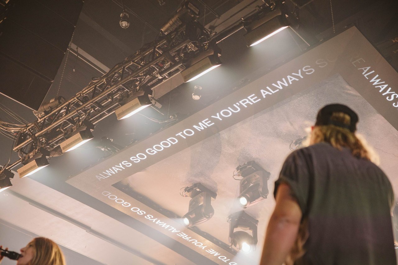 Under hazy stage lights, a capped traveler reads “ALWAYS SO GOOD TO ME” on the ceiling—budget vibes, real atmosphere.