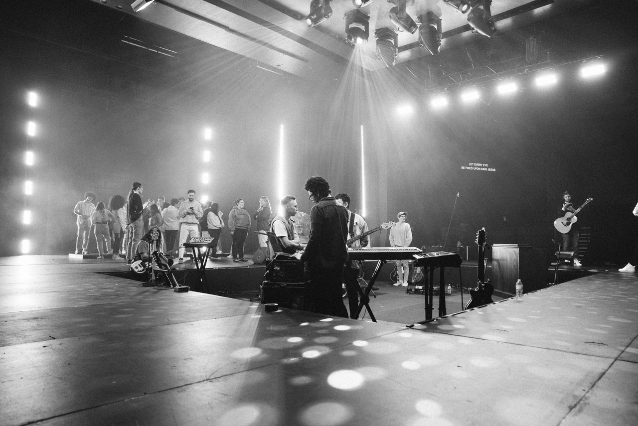 Black-and-white shot of a rehearsal stage: musicians jamming up front, crew hanging in back, gear and cables scattered everywhere.