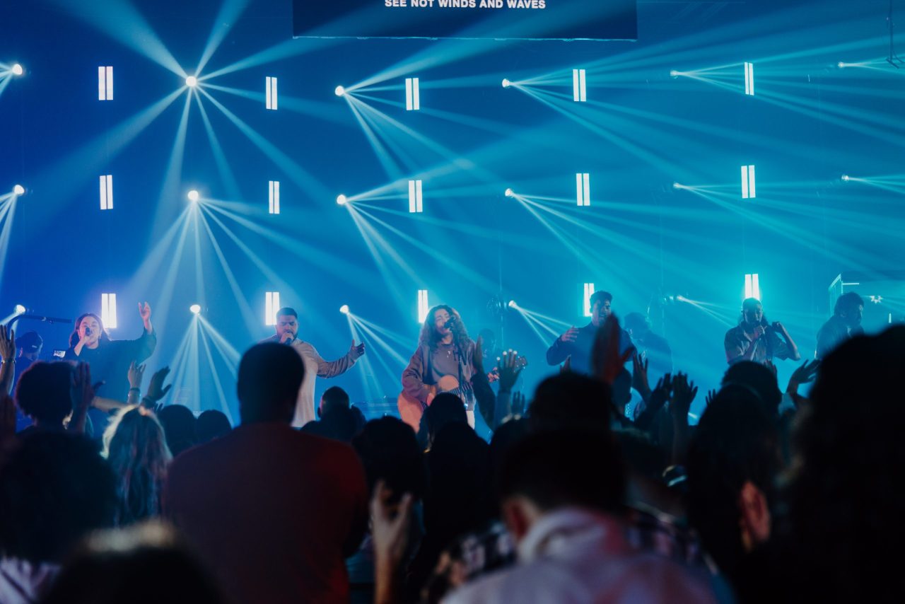A band jams under bright blue lights, six musicians on stage, crowd in front with hands up—buzzing energy, real concert vibes.