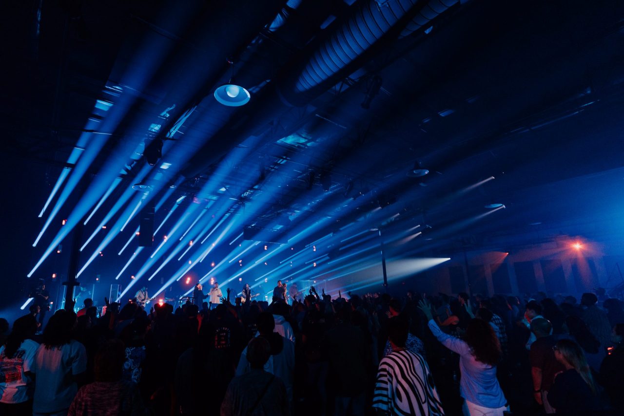 Packed crowd at an indoor concert, blue spotlights cut through haze as people stand close, arms up, soaking in the lively scene.