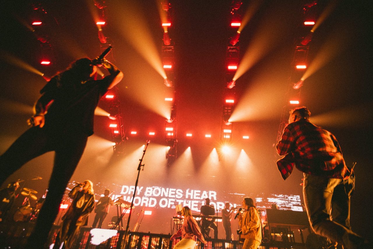 Energetic concert with bold orange lights; two singers fire up the crowd as “DRY BONES HEAR THE WORD OF THE LORD” beams behind them.