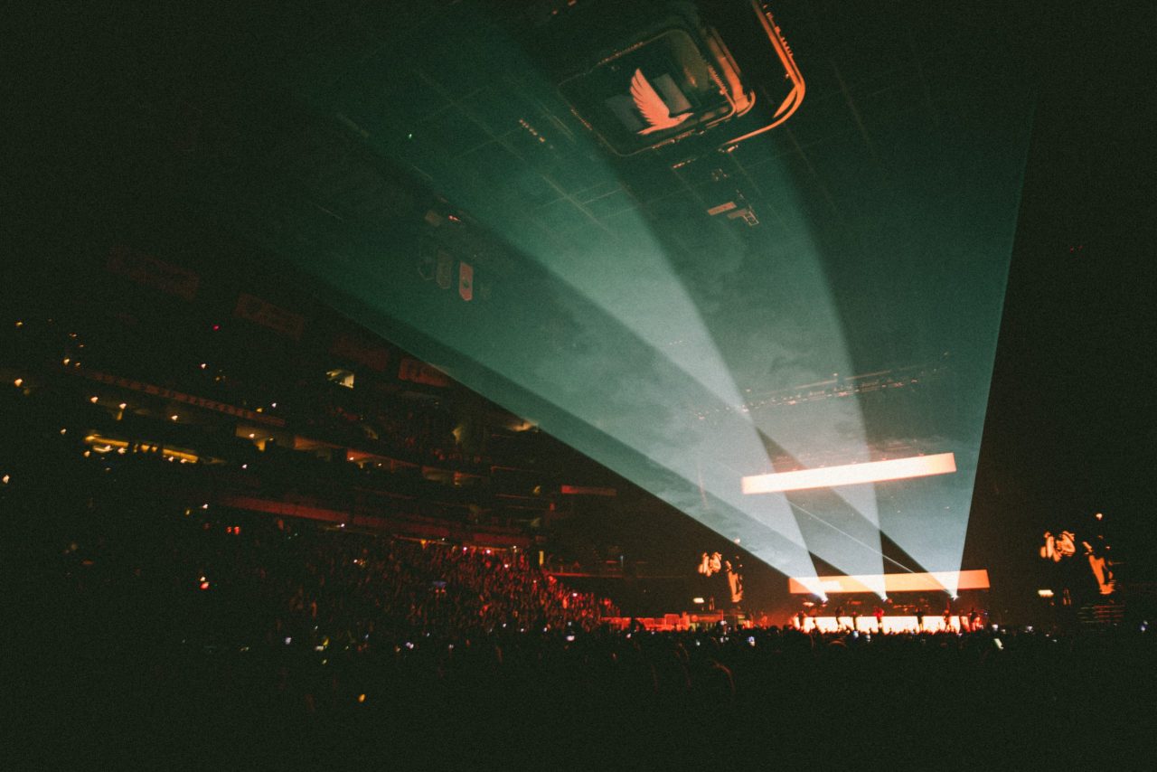 Packed indoor concert arena, bright stage lights cut through haze, crowd mostly in shadow with glowing phones, ceiling beams above.