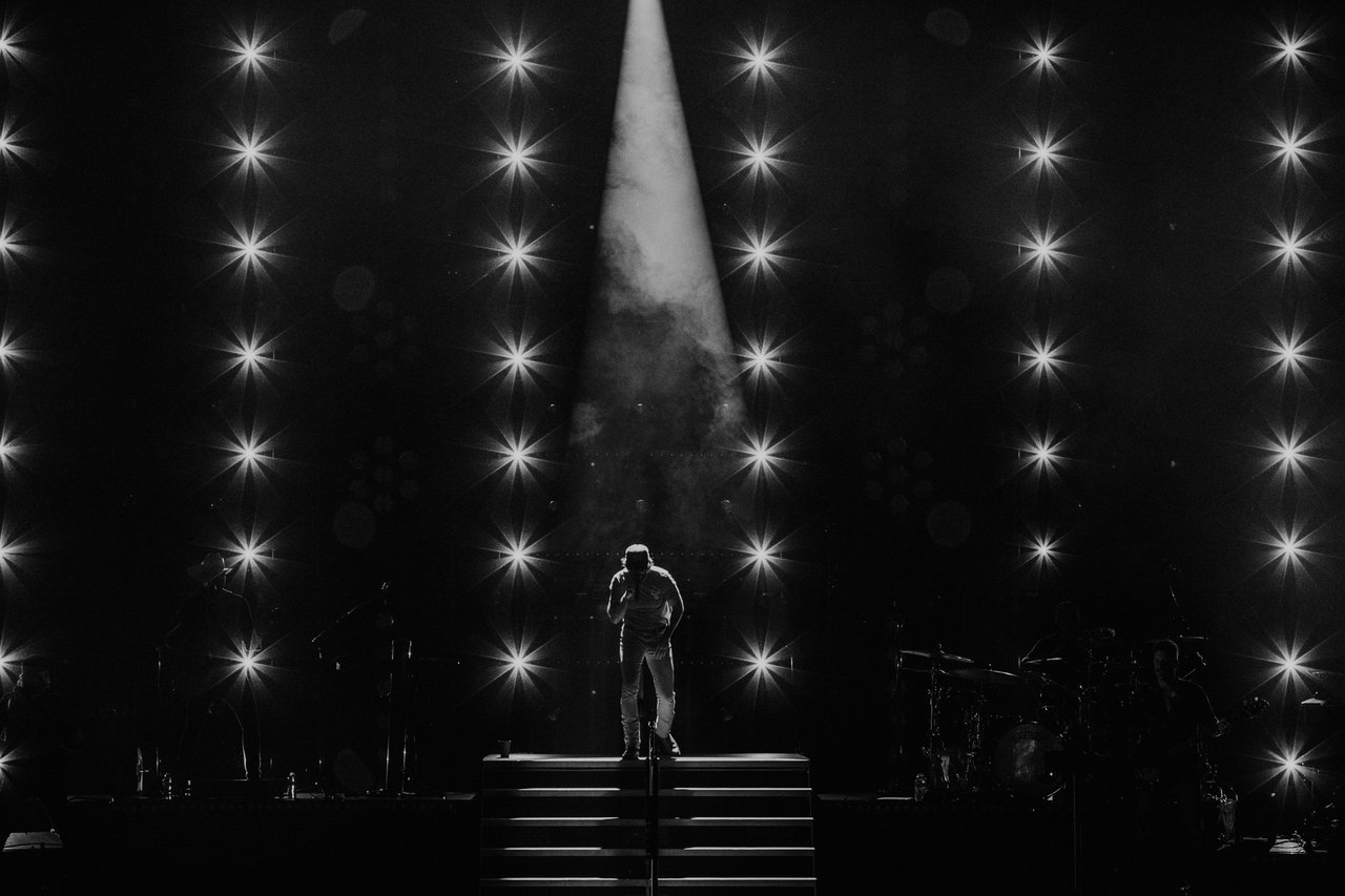 Solo singer in spotlight on a dark stage, starburst lights behind. Musicians in the shadows. Simple, striking, black and white scene.