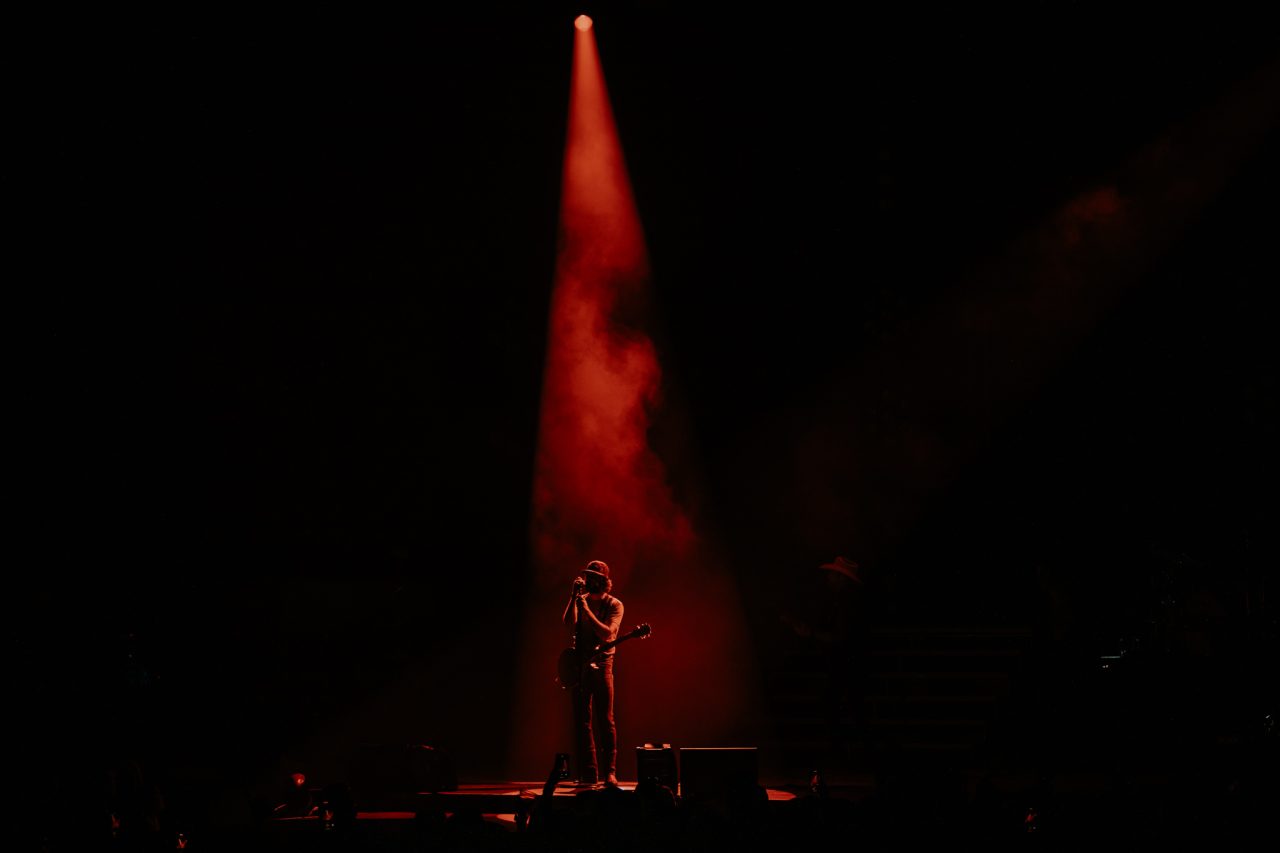 A musician rocks out on stage under a red spotlight, smoke swirling, while the rest of the concert fades into dark and moody vibes.