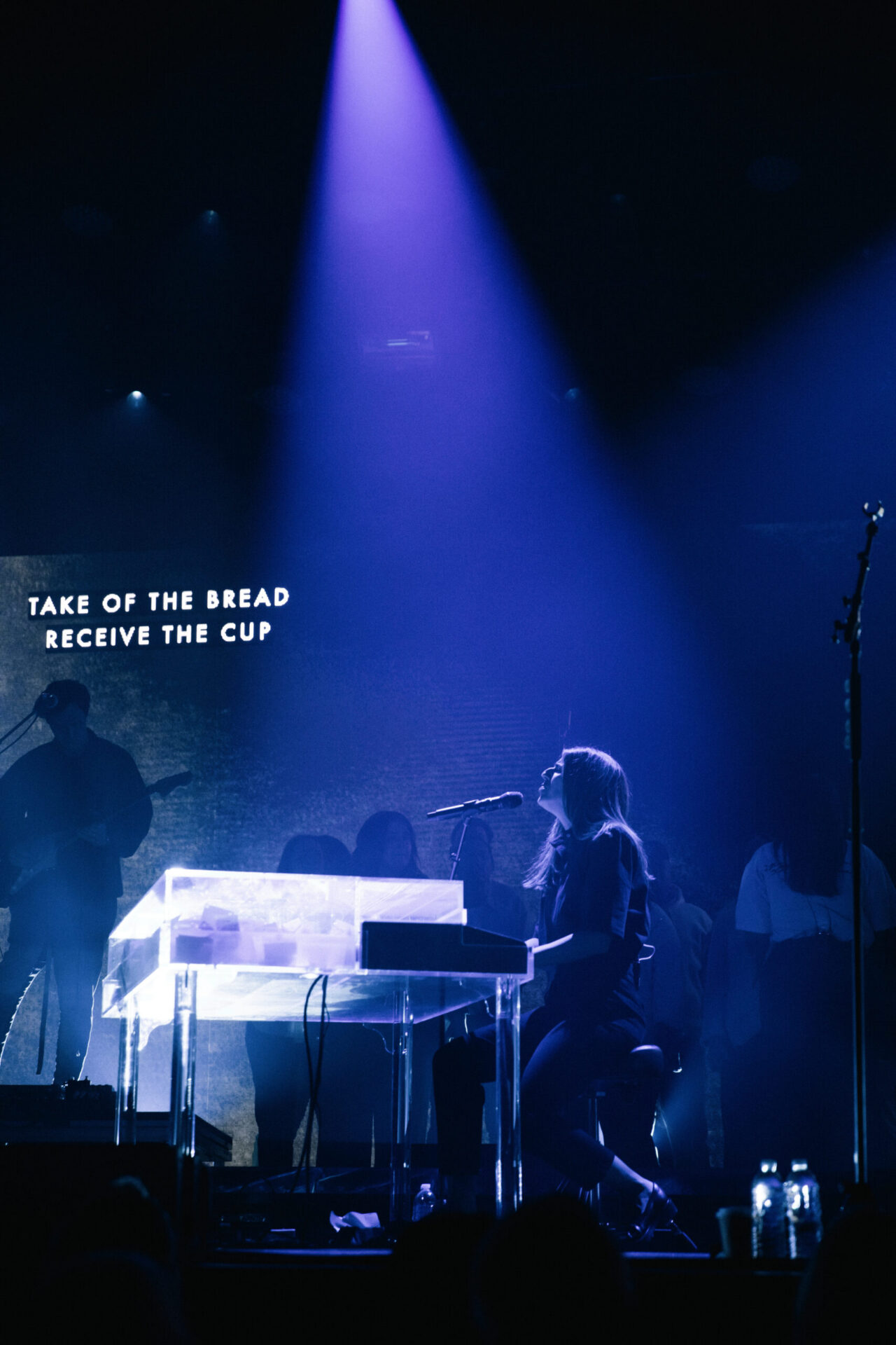 Onstage, a woman at a see-through piano is lit by vivid lights. Behind her, people stand under the sign: TAKE OF THE BREAD RECEIVE THE CUP.