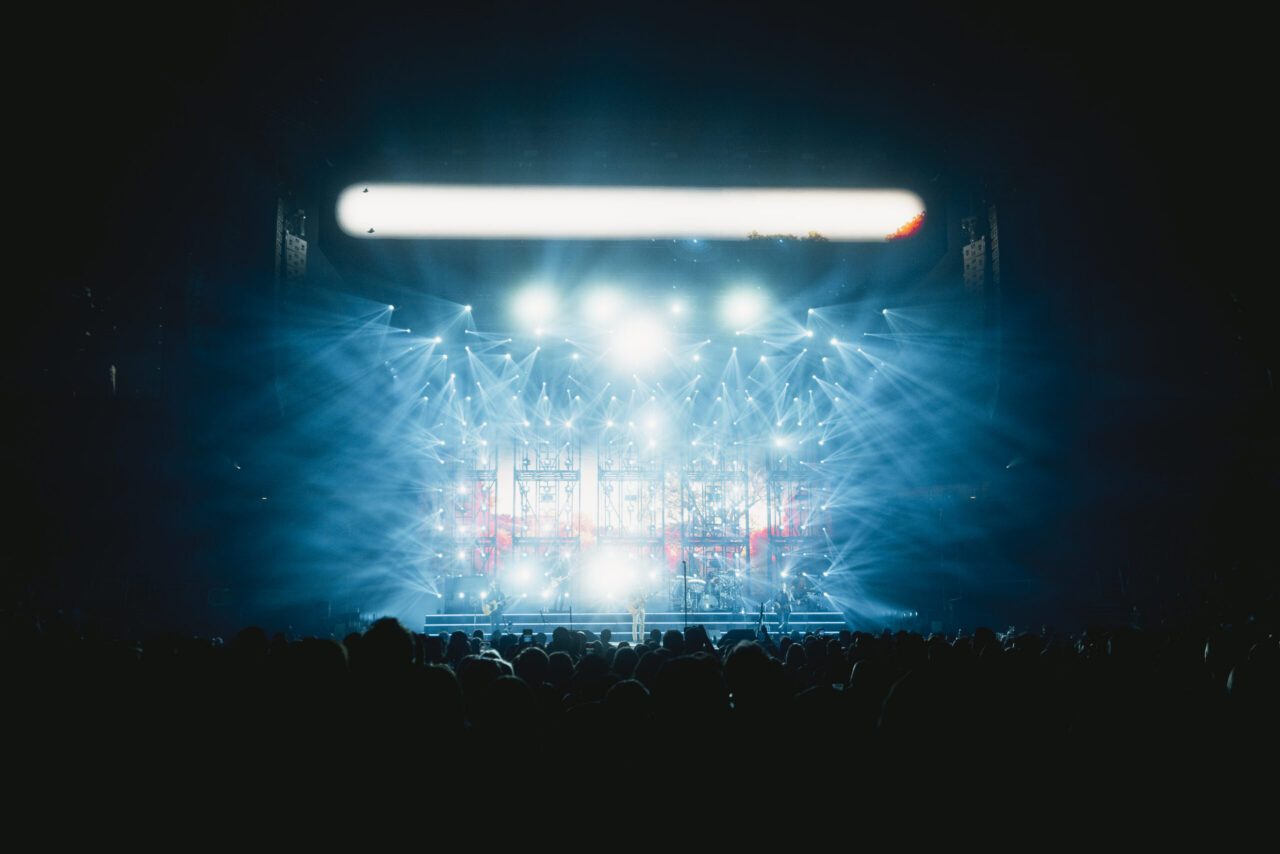 Blue-lit concert stage with bold spotlights, grid of lights and screens; silhouetted crowd upfront. Dramatic, electric vibe.