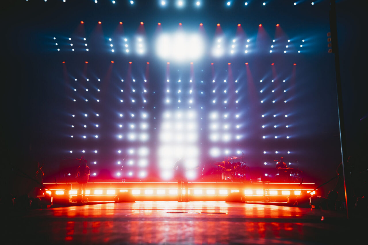 A concert stage pops with blue, white, and red lights. Musicians are silhouetted; the crowd area up front is pure darkness.