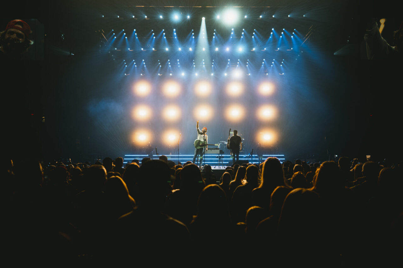 A buzzing crowd watches a musician rocking out center stage under cheap spotlights, backed by glowing lights in a packed, dark venue.