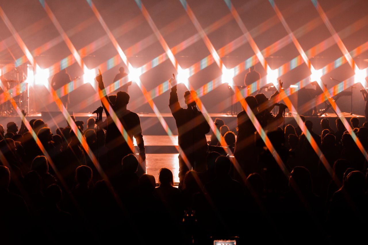 A silhouetted crowd raises their hands at Bayside Christmas 2022 as diagonal streaks of stage light cut across the room.