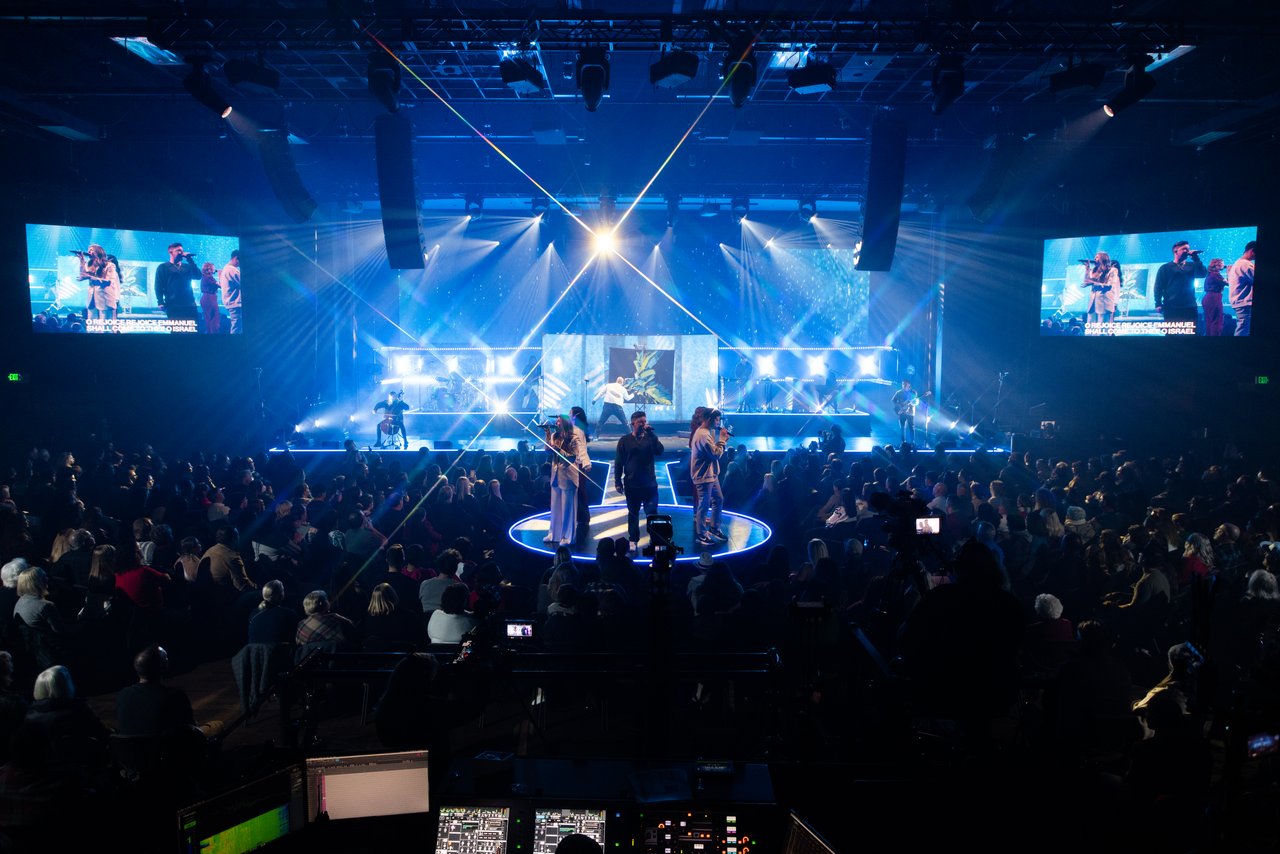 A large audience watches a live music performance on a brightly lit stage, featuring multiple singers and musicians. Two large screens display close-ups, showcasing the stage production at Bayside Christmas 2022.