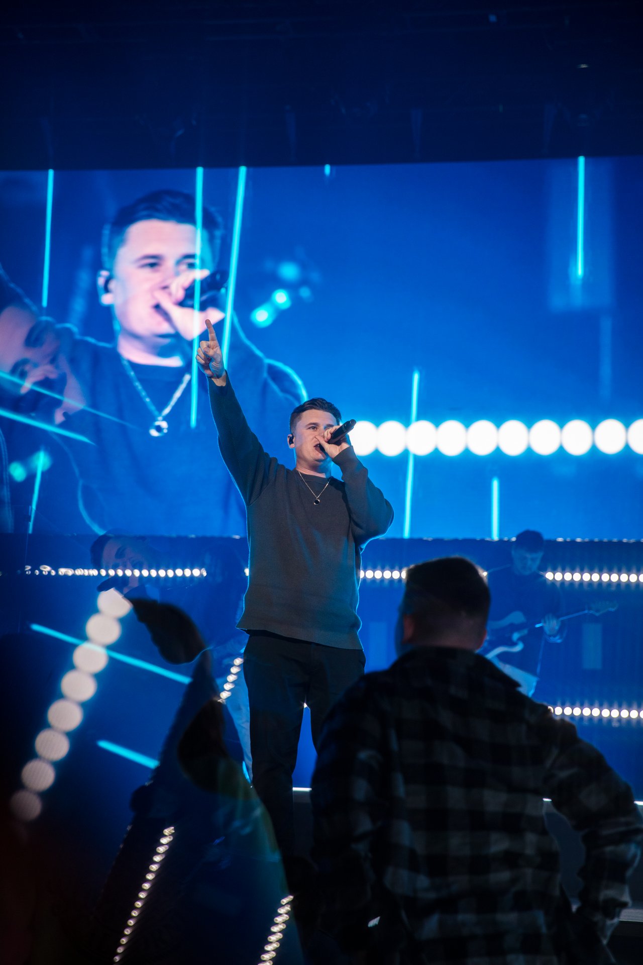 A singer holds a microphone and points upward on stage during Bayside Christmas 2022, with a large screen showing his image, dynamic lighting design, and other musicians in the background.